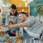 Child is playing with mother and father cheerful adults at home building with construction blocks sitting on floor together. Youth and family concept.